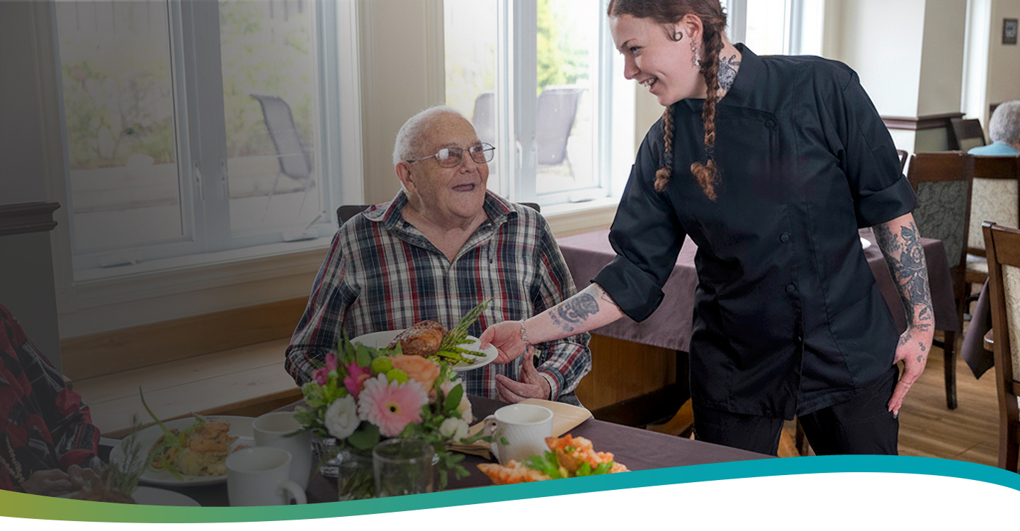 Server presenting a beautifully plated dinner to a smiling resident in the bright dining room at Atrium Retirement Residence in Orillia.